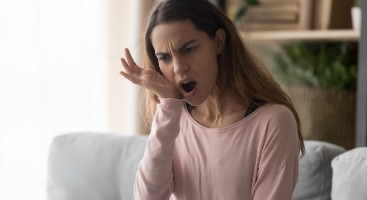 Woman sitting on couch and holding the side of her jaw in pain